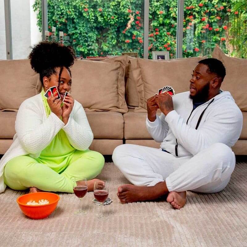casal sentados em uma sala, por cima de um tapete, com uma bacia de pipoca e duas taças de vinho, os dois estão com um baralho de uno jogando.