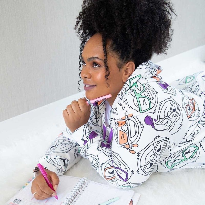 mulher deitada, com duas canetas, uma em cada mão, a mão esquerda apoiada no queixo, e mão direita sobre um caderno escrevendo.