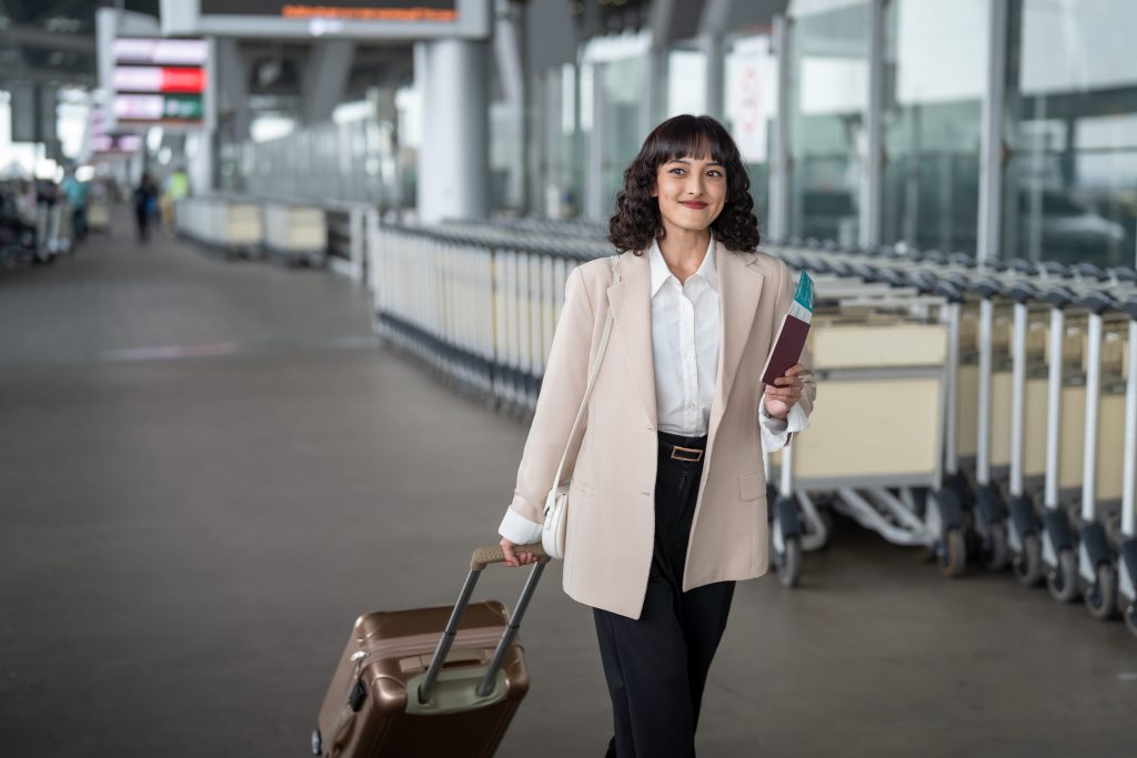 Mulher de casaco bege, sorrindo e carregando uma mala de rodinhas em um aeroporto.