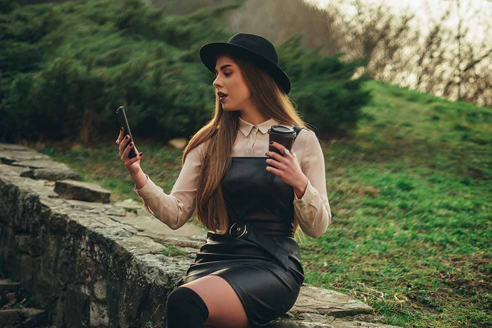 estilo sexy mulher sentada em um pequeno muro de pedras com um vestido preto em couro e uma camisa bege por baixo, junto de um chapeu preto e botas pretas com um celular em mãos