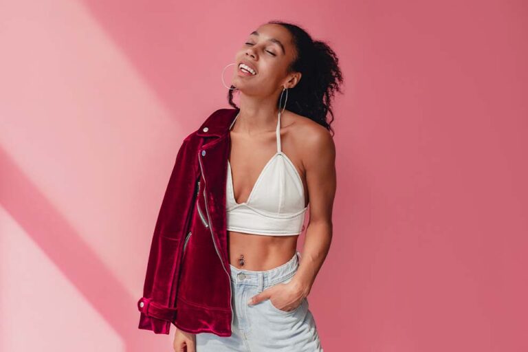 Mulher com um top branco, jaqueta de veludo vermelha sobre o ombro e piercing no umbigo, em um fundo rosa.