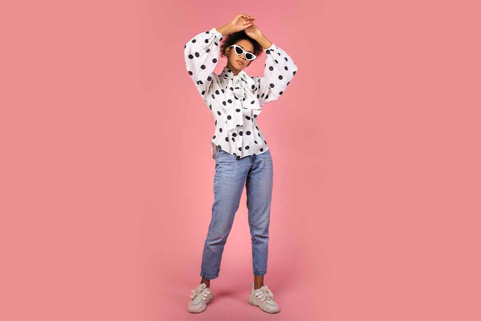 mulher usando uma bata e calça jeans junto de um tênis branco em um fundo rosa claro