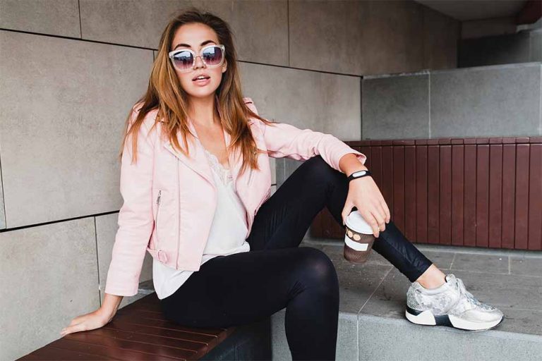 mulher usando uma camisa branca com uma jaqueta rosa claro e uma calça preta e tênis sentada em uma escada de concreto