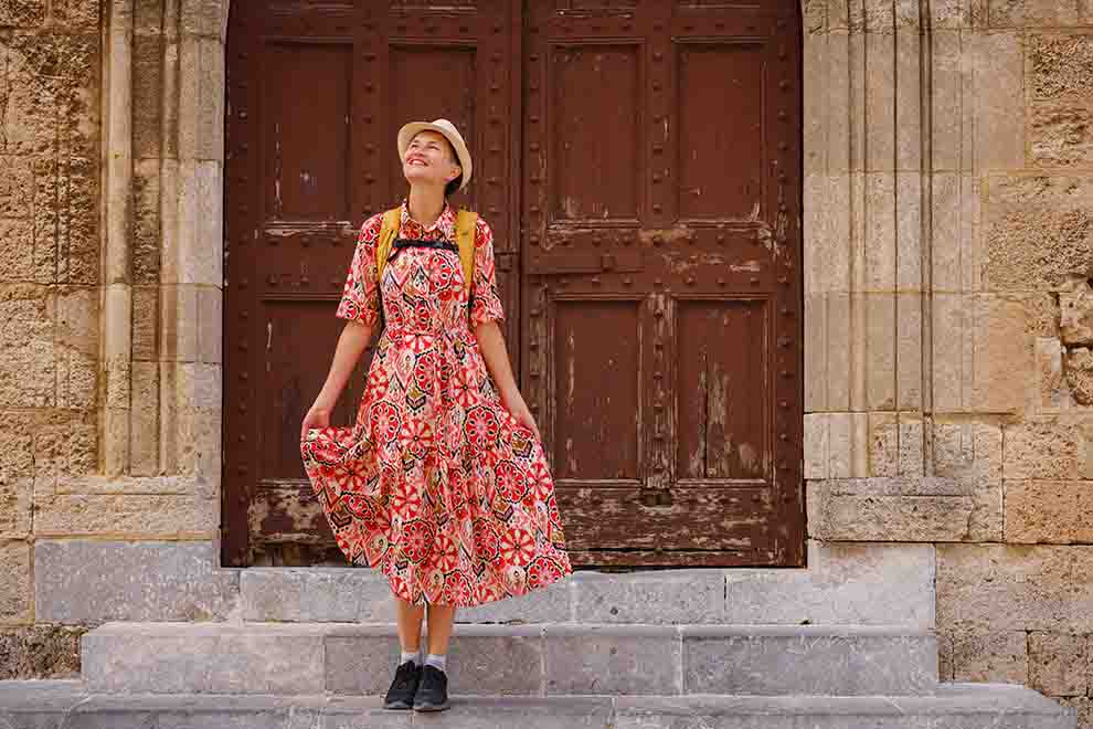 Mulher sorridente em vestido florido diante de porta antiga.