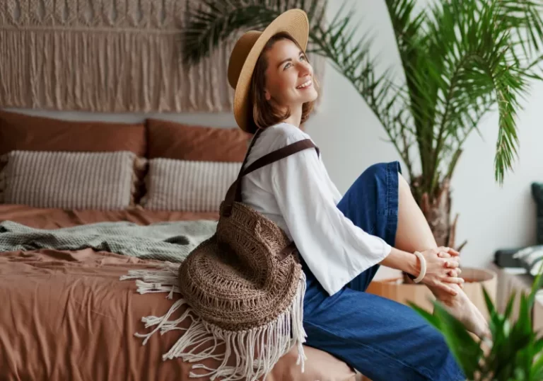 mulher sorrindo sentada na cama com chapéu e bolsa de lado
