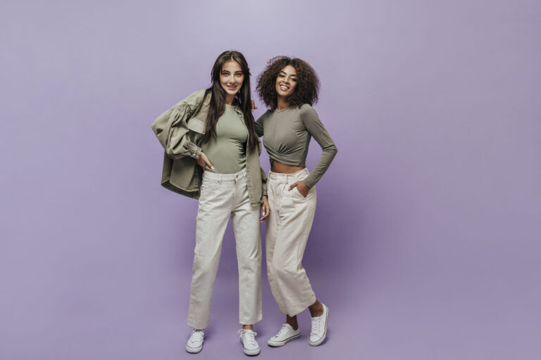 Imagem com uma dupla de modelos femininas vestindo calças cor caqui e blusa manga longa cor verde-escuro no fundo roxo