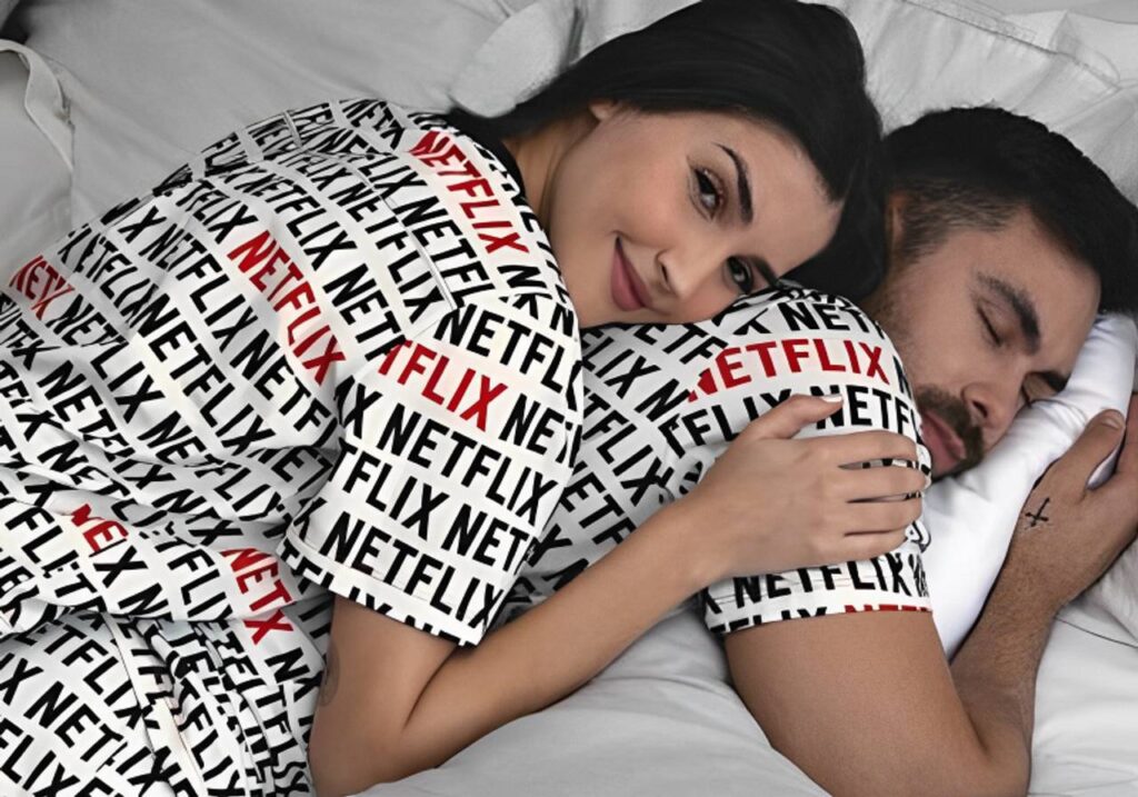 pijamas de casal combiando casal deitados homem dormindo de bruços e mulher em cima dele com pijamas combinando