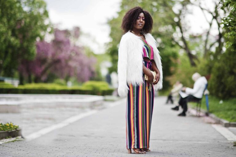 Imagem de uma modelo feminina, vestindo um conjunto de roupas confortáveis, com uma calça estampada de listras verticais, com um casaco branco, em pé, com o fundo de parque de dia.