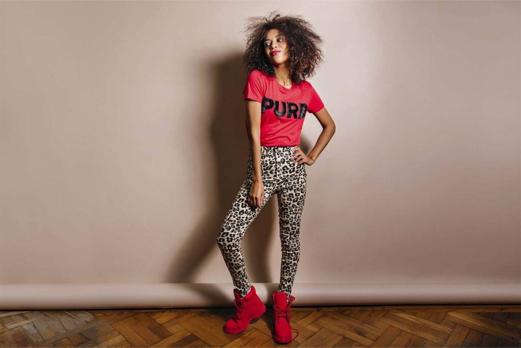 modelo calca estampada amarelo Imagem de uma modelo feminina, em pé, vestindo uma camiseta vermelha, bota vermelha e calça estampada de onça, em um fundo bege de estúdio fotográfico.