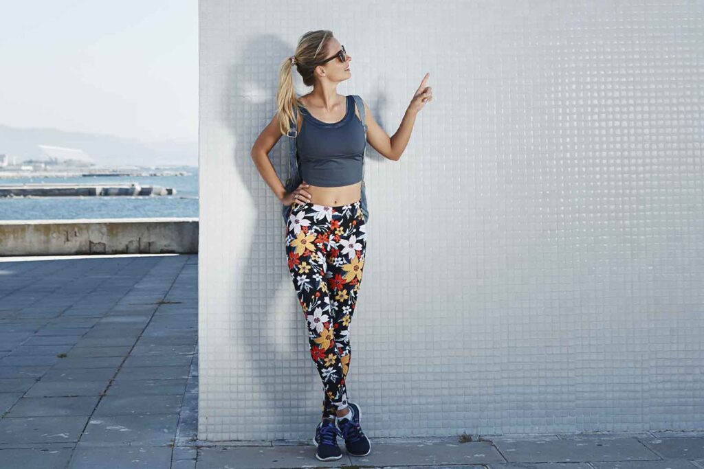 modelo calca estampada colorida Imagem de uma modelo feminina, em pé, vestindo uma calça estampada florida, top cinza, tênis, óculos escuros e uma mochila nas costas, com o fundo de parede cinza, em meio urbano, com um pedaço do mar ao fundo e de dia.